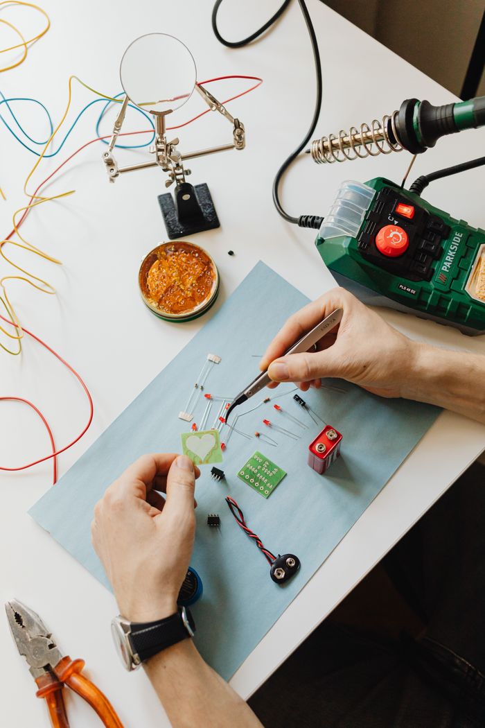 Hands assembling electronic components on a work table with tools and wires.